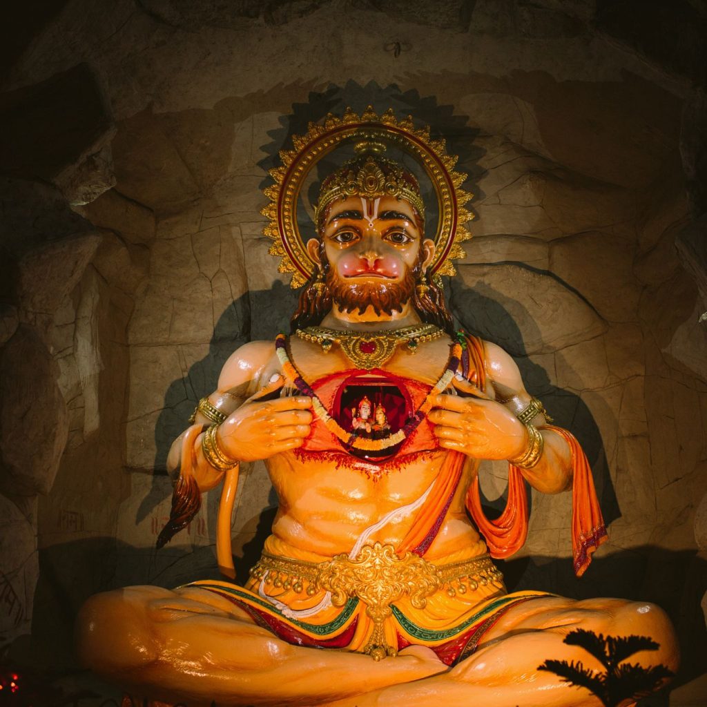 A grand and magnificent statue of Lord Hanuman, tearing open his chest to reveal images of Shri Ram and Sita, with a cave-like wall in the background.