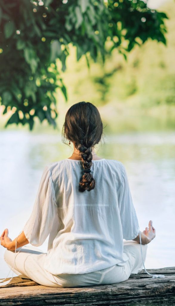 A woman in a white outfit sits by a river, meditating with her hair tied in a braid, under the shade of green tree leaves.