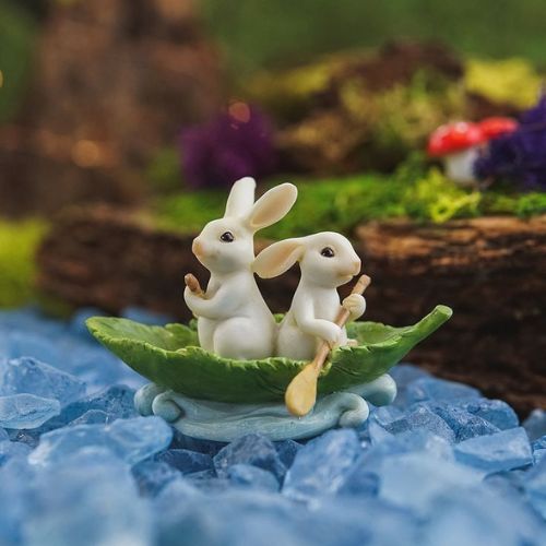 Two white rabbits sitting in a small green boat, paddling on a river made of blue stones, with a fairy-tale forest background.