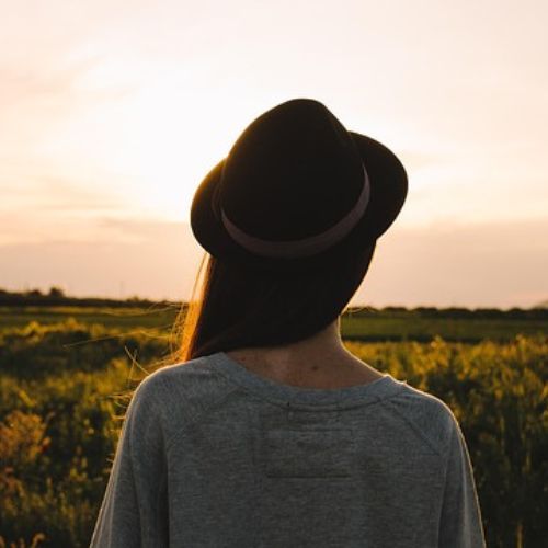 A woman in a gray sweater and black hat stands in a field at sunset, facing away from the camera, watching the golden sky as the sun sets on the horizon.