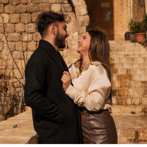 A couple laughing together near a rustic stone staircase, sharing an intimate and joyful moment in warm sunlight.