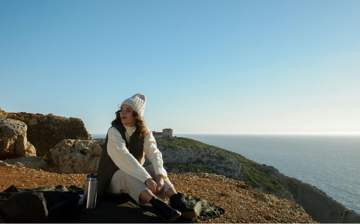 A woman sitting on rocky cliffs near the sea, dressed in warm white attire, enjoying the natural scenery with a thermos beside her.