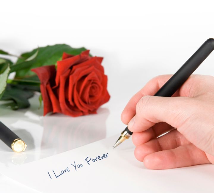 A person is writing "I Love You Forever" on white paper with a black pen, with a red rose placed beside it.