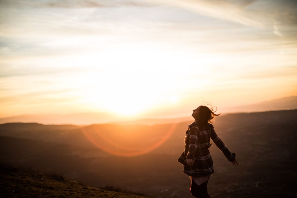 A young woman in a white polka dot dress stands in a golden field at sunset, spreading her arms joyfully. The warm glow of the setting sun creates a peaceful and serene atmosphere.