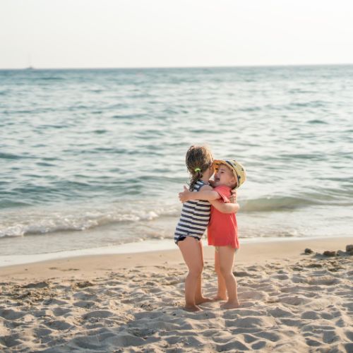 Two young children hugging on a sandy beach with the ocean in the background, representing friendship and warmth.