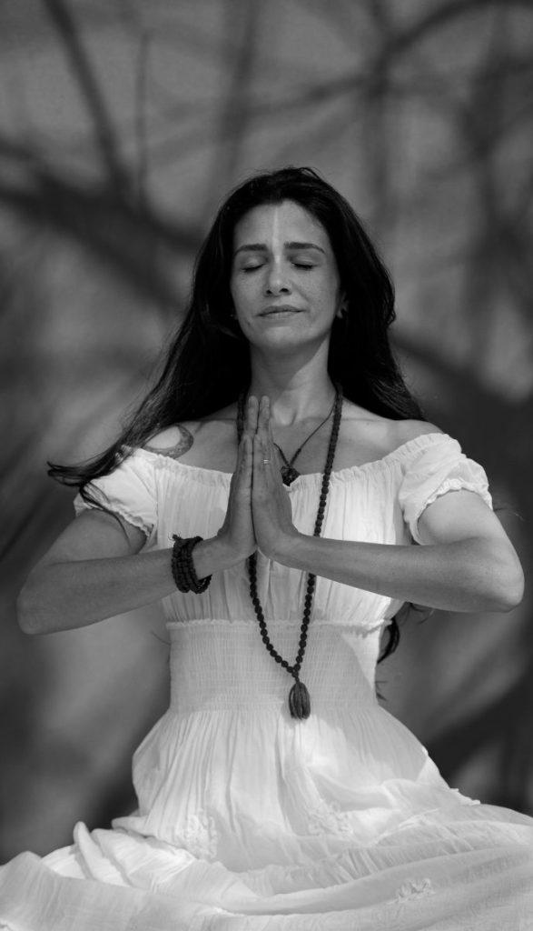 A black-and-white photograph of a woman in a white dress meditating with her hands pressed together in a prayer position. She has long, dark hair, closed eyes, and a serene expression. Shadows of tree branches are visible on the background wall.