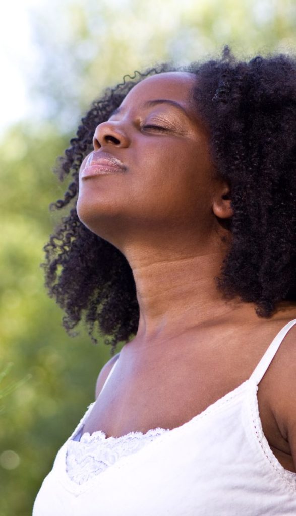 A woman with curly black hair tilts her head back and closes her eyes, basking in the sunlight. She wears a white spaghetti-strap dress and stands in an outdoor setting with blurred green foliage in the background.