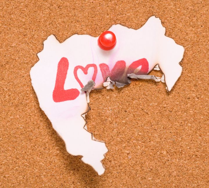 A torn and slightly burned piece of paper pinned to a corkboard with a red pushpin, featuring the word 'LOVE' written in bold red letters, with the 'O' replaced by a heart shape.