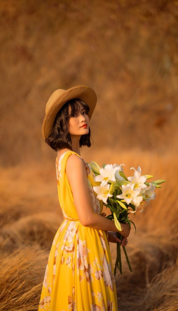 A woman in a yellow floral dress stands in an open field, holding a bouquet of white lilies, glowing in the sunlight.