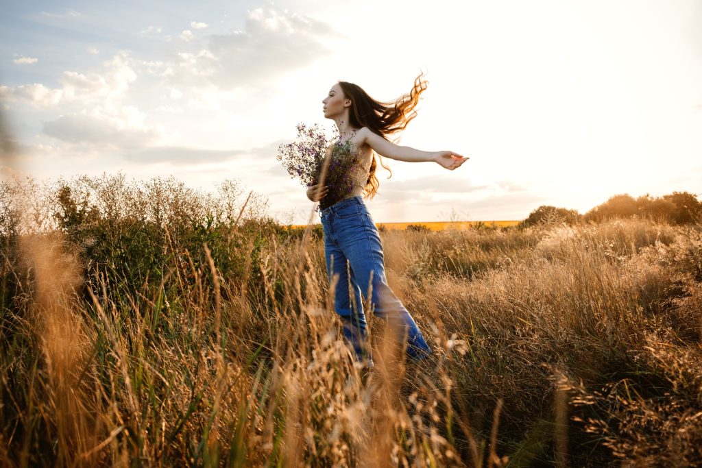 A young woman with long, flowing hair dances freely in a sunlit field, holding a bouquet of wildflowers. She wears blue jeans and a floral top, embracing the beauty of nature as the wind moves through the tall grass.