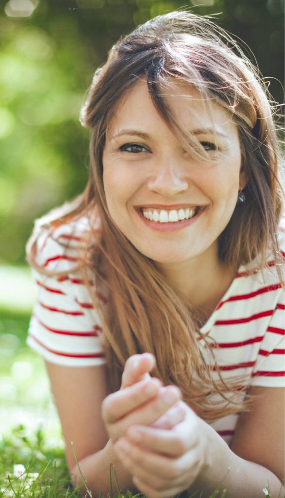 A smiling woman in a red and white striped shirt lies on green grass, looking happily at the camera.