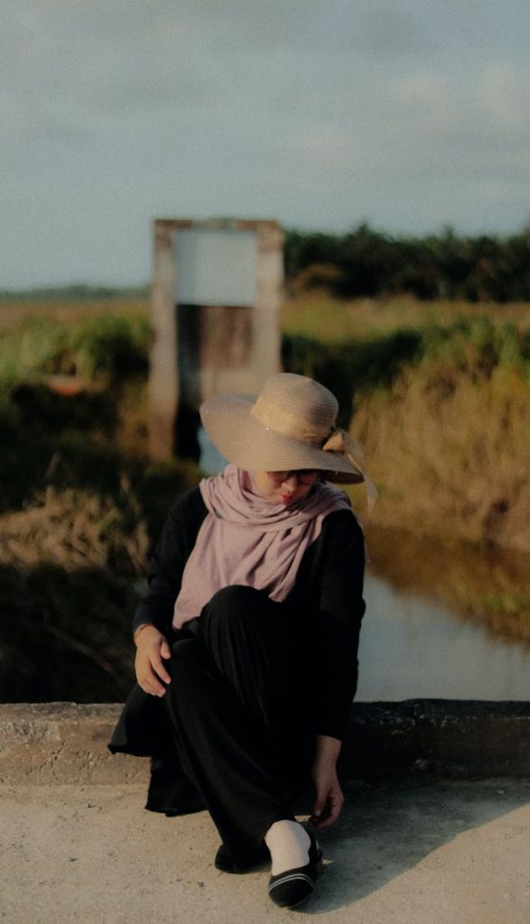 A woman wearing a wide-brimmed hat and a scarf sits on a concrete ledge near a water body, with a blurred rural landscape in the background.