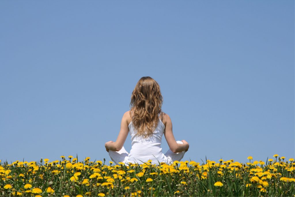 Woman meditating in a field of yellow flowers under a clear blue sky.