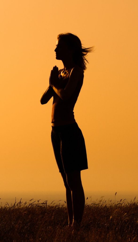 A silhouette of a woman practicing meditation or prayer in a grassy field during sunset, with an orange sky in the background.