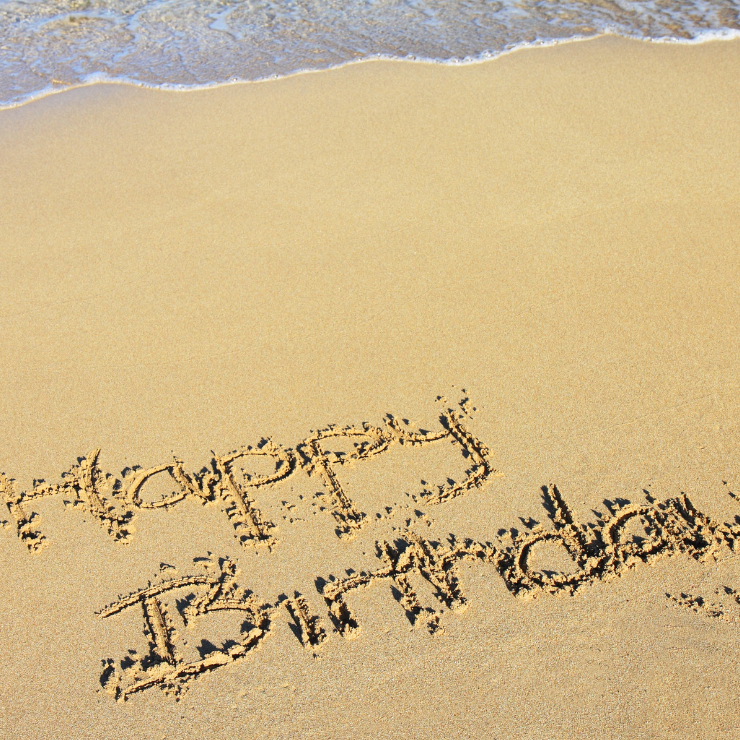 Happy Birthday' written in the sand on a beach with gentle ocean waves approaching.
