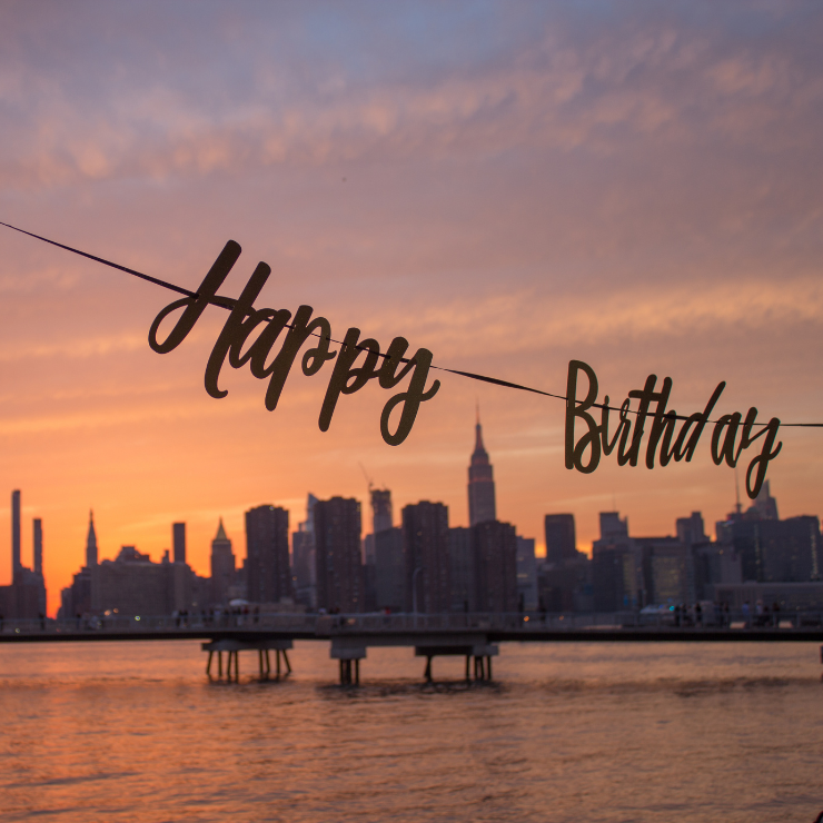 A "Happy Birthday" banner hangs against a sunset-lit city skyline, with the silhouette of skyscrapers in the background.