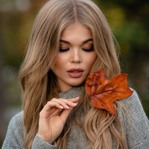 A beautiful blonde woman smiles while holding a vibrant autumn leaf against a blurred natural background
