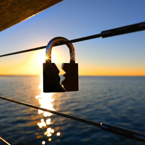 A broken padlock rests on a railing, with a scenic view of the ocean in the background