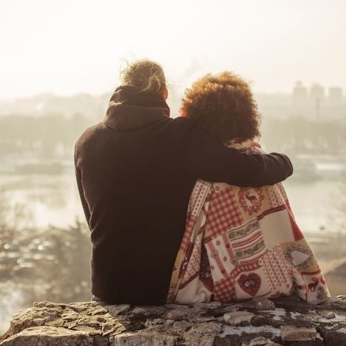 A couple sits on a rock, gazing at a sprawling cityscape under a clear blue sky