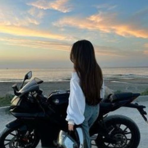 A girl stands beside a motorcycle on a sandy beach, with waves gently crashing in the background