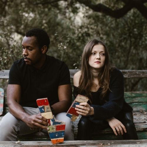 A man and woman sit on a bench holding a box together, smiling at each other in a park setting