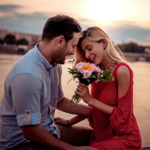 A man and woman sit on the ground, each holding a bouquet of colorful flowers, smiling at each other