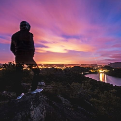 A man stands on a rock, gazing at a serene lake during a vibrant sunset