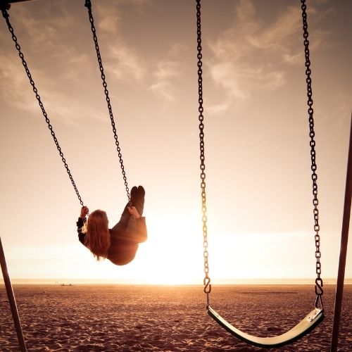 A person swings joyfully on a swing set at the beach with sand and ocean waves in the background