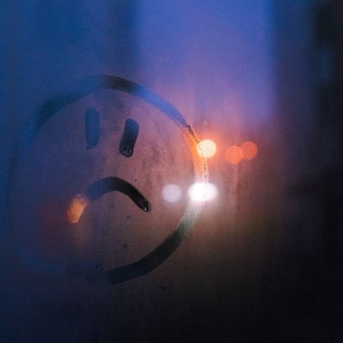 A sad face drawn on a foggy window with a blurred background creating a melancholic atmosphere
