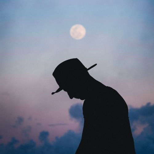 A silhouette of a man in a hat stands against a bright full moon