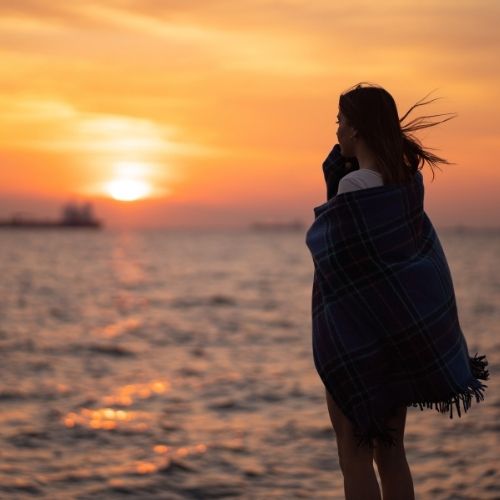 A woman in a blanket stands on the beach, gazing at the vibrant sunset over the ocean