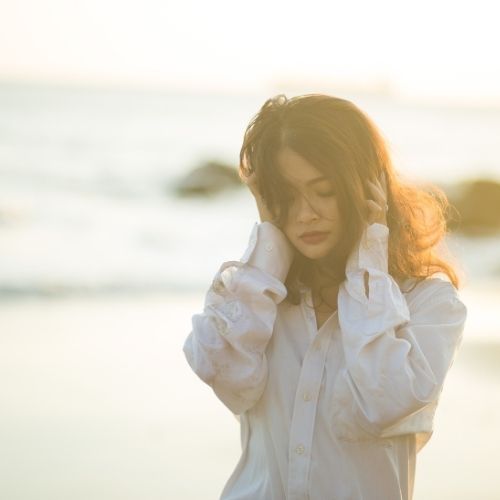 A woman in a white shirt stands on the beach, gazing at the ocean under a clear blue sky