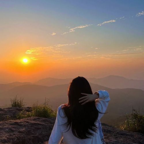 A woman sits on a rock, gazing at a vibrant sunset over the horizon