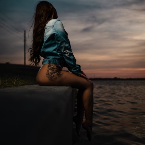 A woman sits on a wall, facing away from the camera, with her hair flowing in the breeze