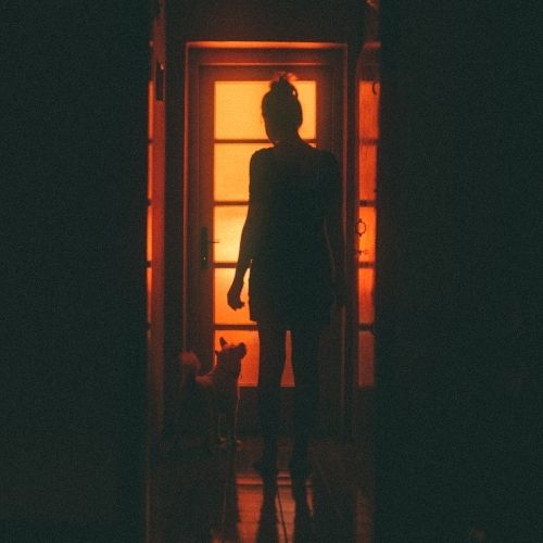 A woman stands in front of a closed door at night, illuminated by soft light, creating a mysterious atmosphere