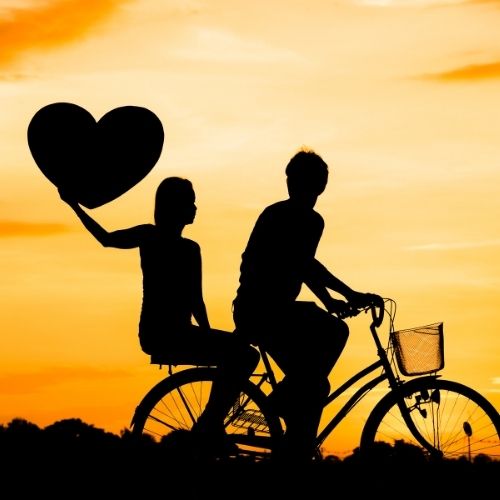 Silhouette of a couple riding a bicycle forming a heart shape with their bodies against a sunset backdrop