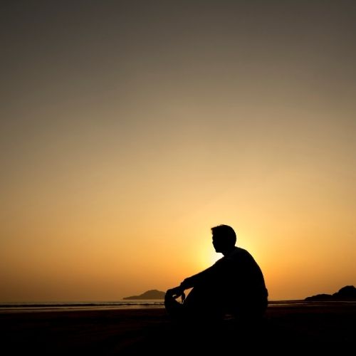 Silhouette of a man sitting on a beach, gazing at a vibrant sunset over the ocean horizon