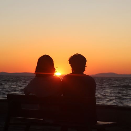 Two people sit on a bench, silhouetted against a vibrant sunset sky