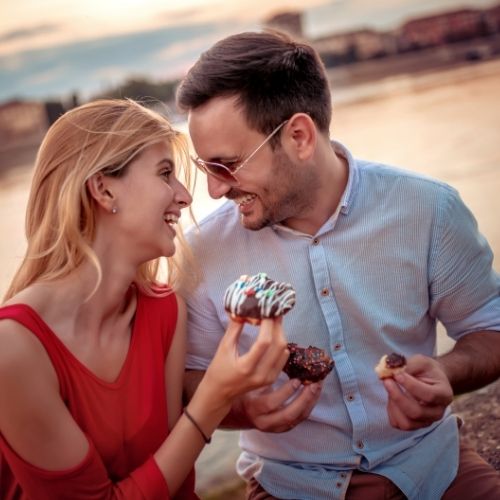 A couple enjoying doughnuts together by a serene river, surrounded by greenery and a peaceful atmosphere