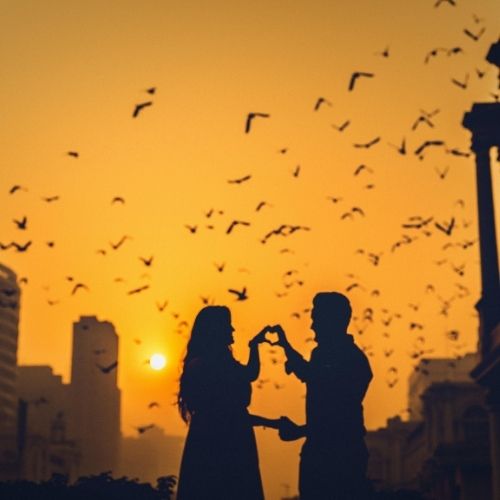 A couple in silhouette, hand in hand, framed by a colorful sunset sky