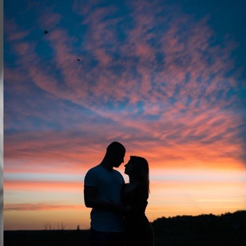 A couple sharing a kiss against a vibrant sunset backdrop, with warm hues of orange and pink illuminating the scene