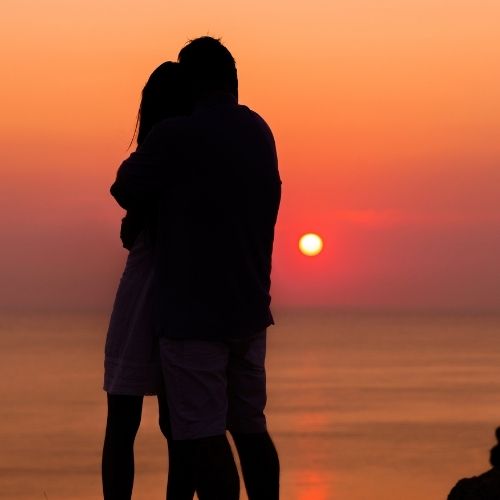 A couple stands on a rock, gazing at the ocean during a vibrant sunset, with warm colors reflecting on the water