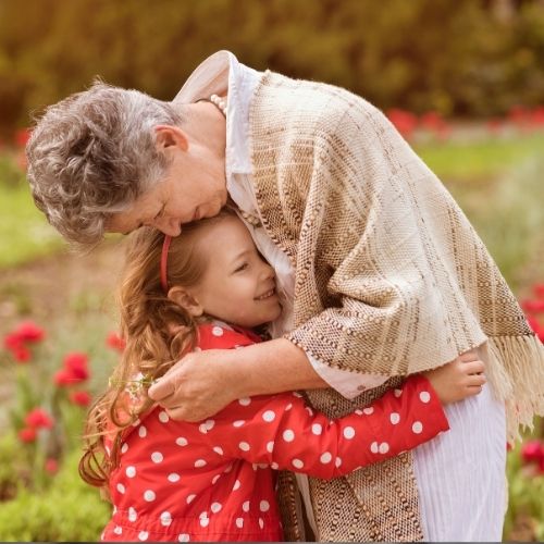 A grandmother and her granddaughter smiling together in a vibrant garden filled with flowers and greenery.