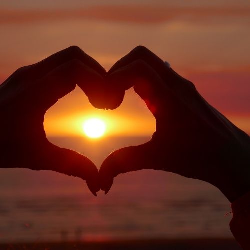 A heart shape created by two hands with a glowing sun in the background, representing affection and positivity