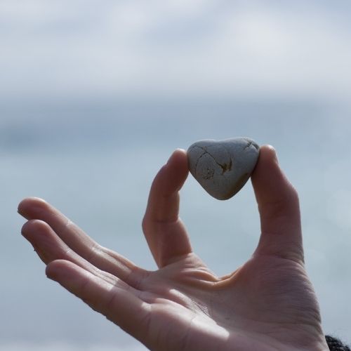 A person stands by the ocean, holding a stone up to eye level, gazing thoughtfully at the water