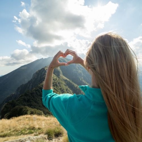 A woman forms a heart shape with her hands while standing on a mountain peak against a clear blue sky