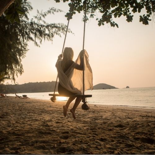 A woman swings joyfully on a swing set at the beach, with the ocean waves and sandy shore in the background