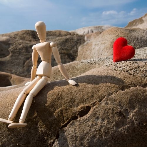 A wooden mannequin sits on a rock, holding a red heart, symbolizing love and creativity in a serene outdoor setting