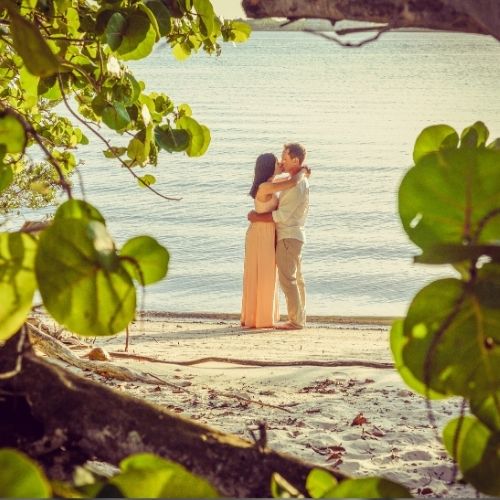 Couple sharing a romantic kiss on a sandy beach during sunset, with gentle waves in the background