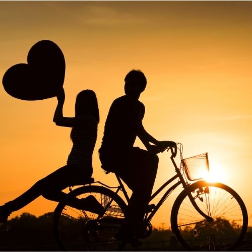 Silhouette of a couple riding a bicycle, forming a heart shape with their bodies against a sunset backdrop